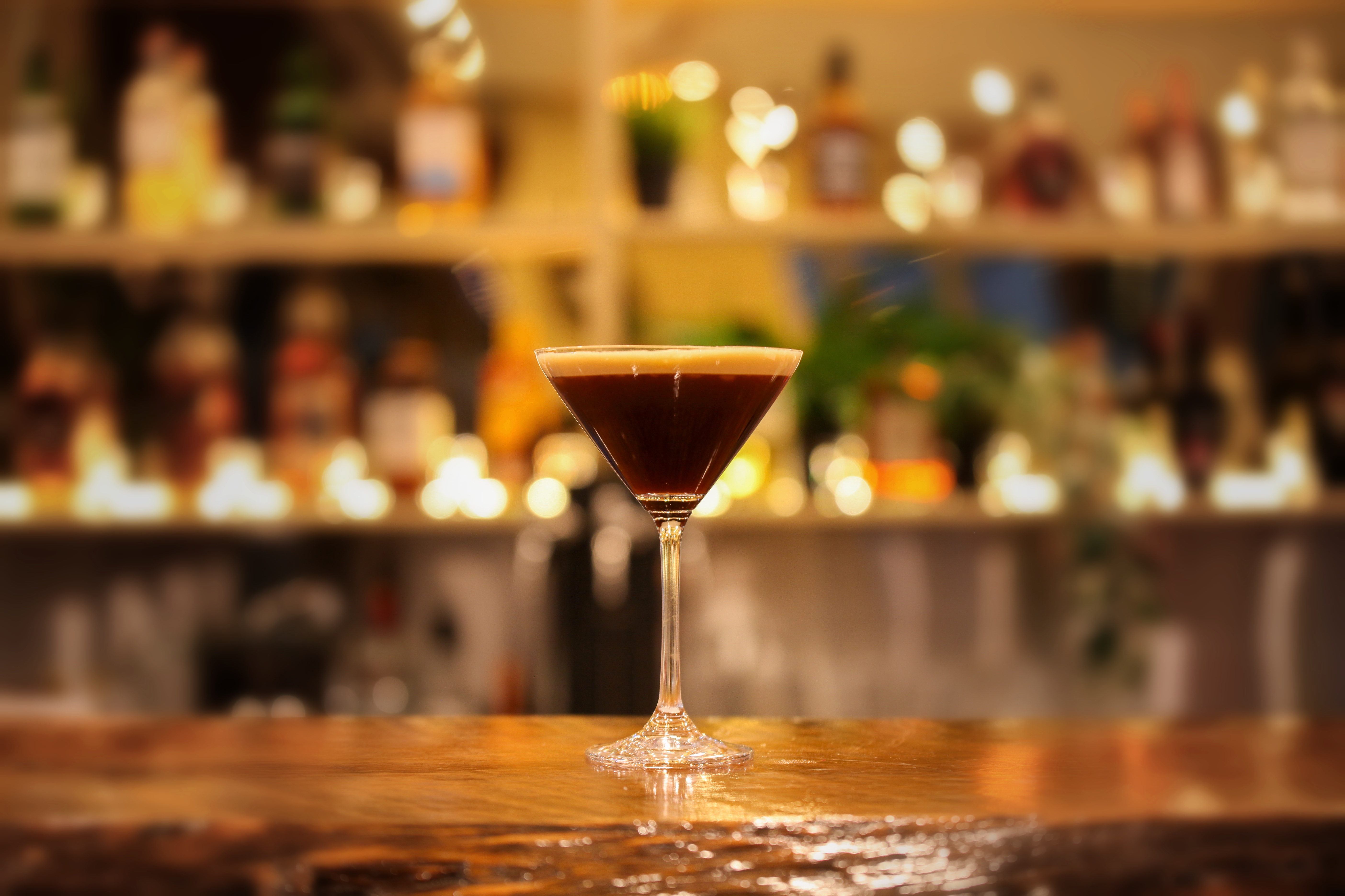 Virgin espresso martini in a martini glass with a creamy crema top on a timber bar, glowing bar shelves behind