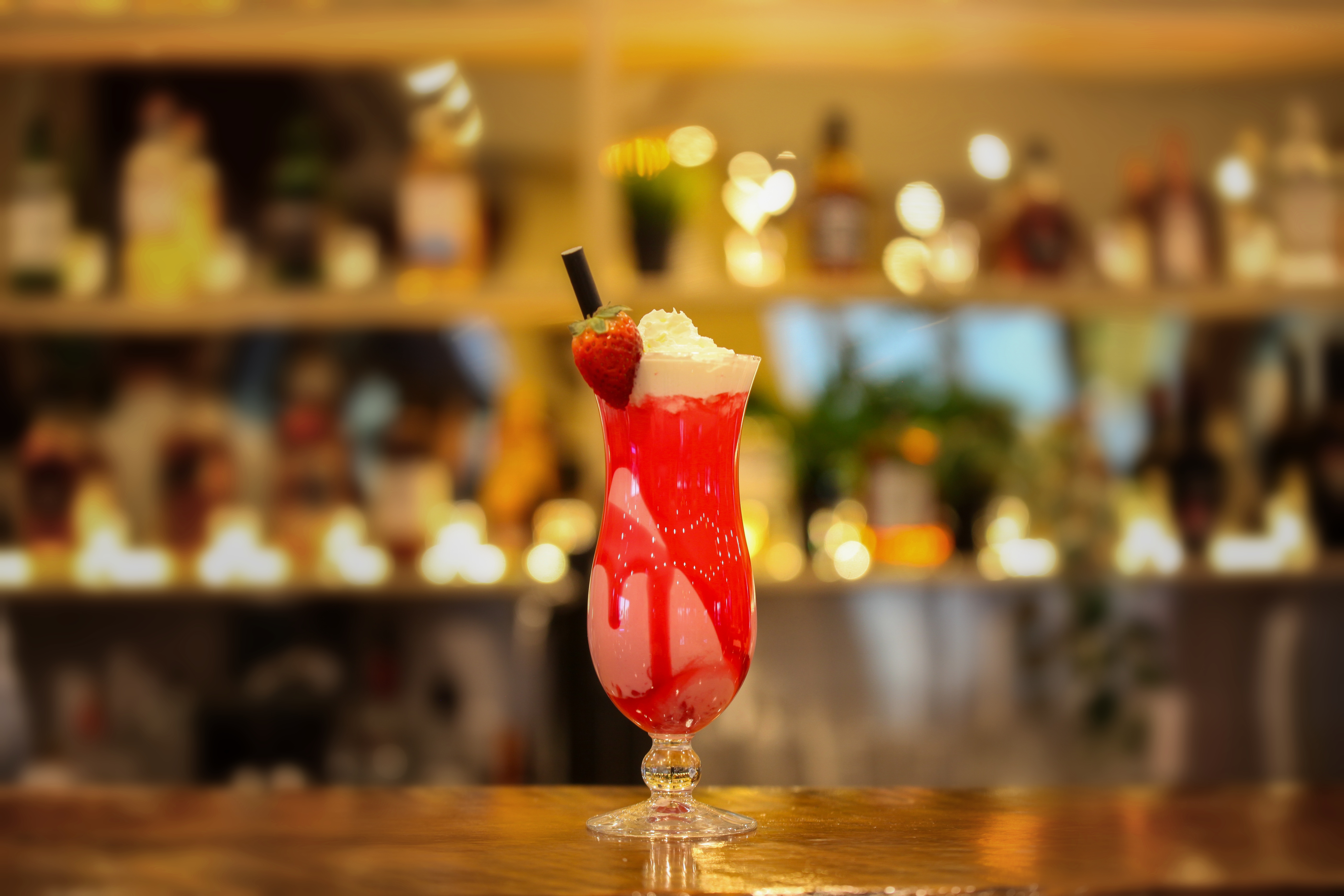 Strawberry shake in a hurricane glass with strawberry sauce, whipped cream and a fresh strawberry, glowing bar shelves behind