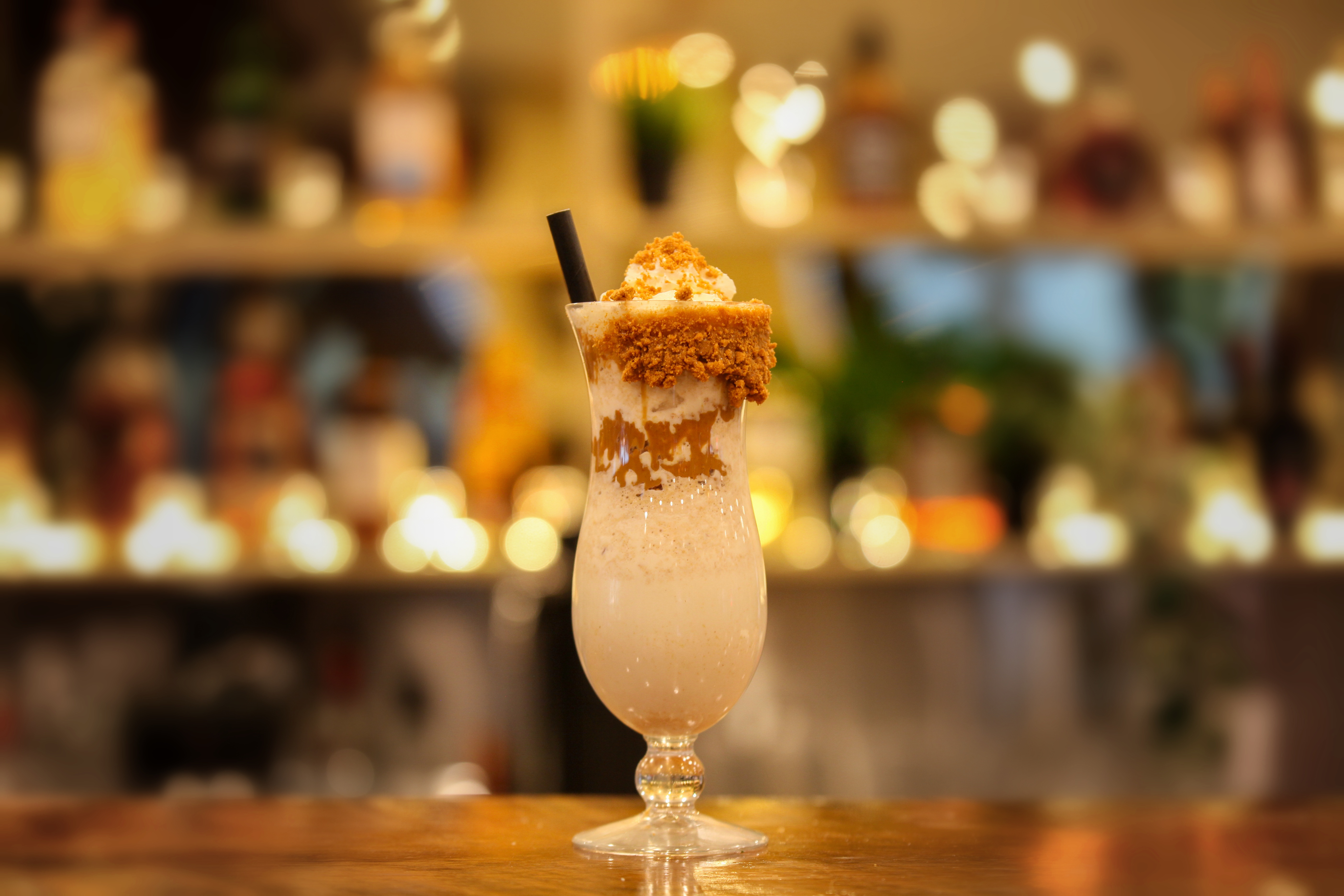 Loaded Biscoff and banana shake in a hurricane glass with a biscuit crumb rim, caramel drizzle, whipped cream and a black straw, glowing bar shelves behind