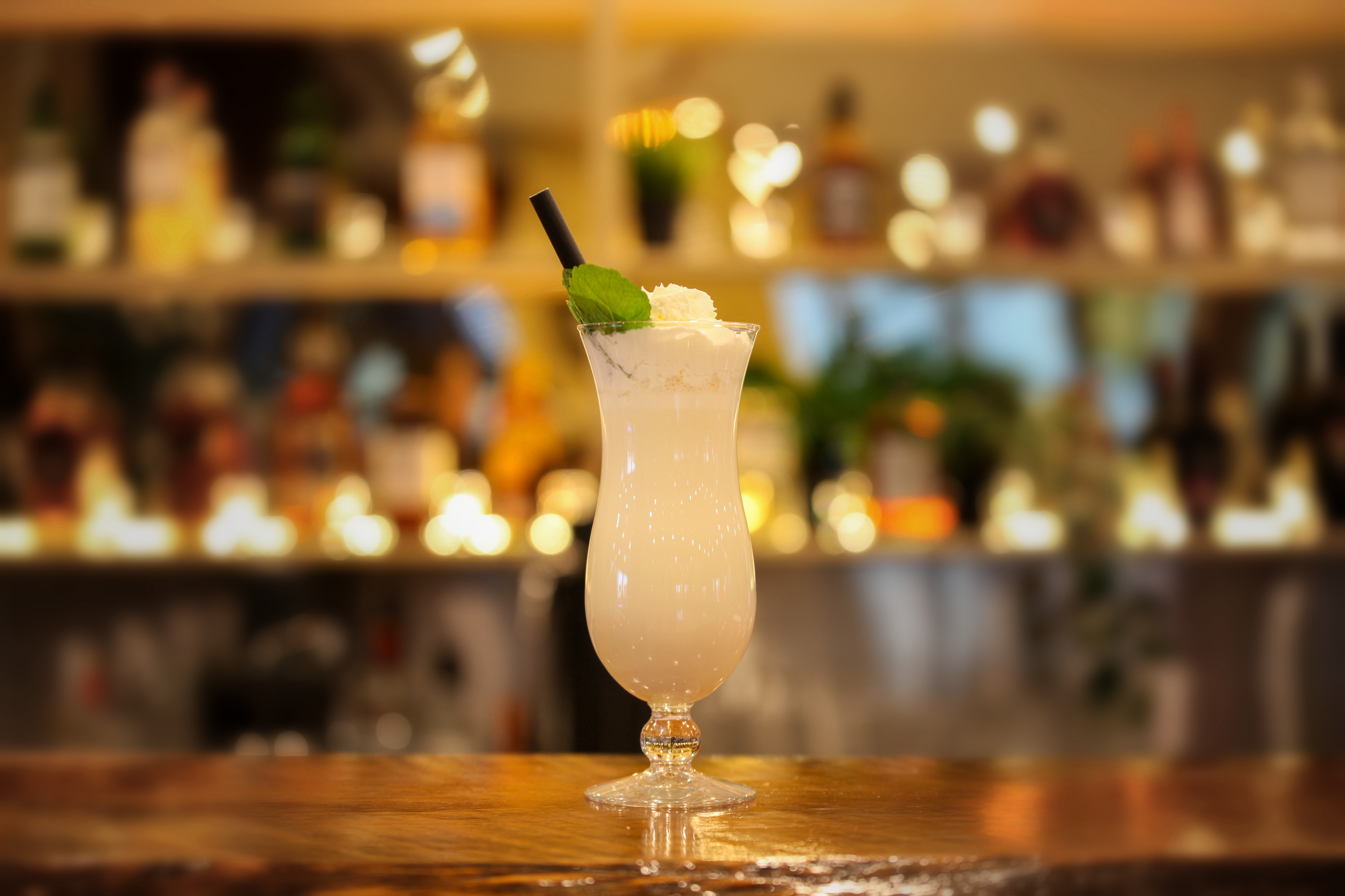 Creamy white shake in a hurricane glass topped with whipped cream and a fresh mint leaf, glowing bar shelves behind