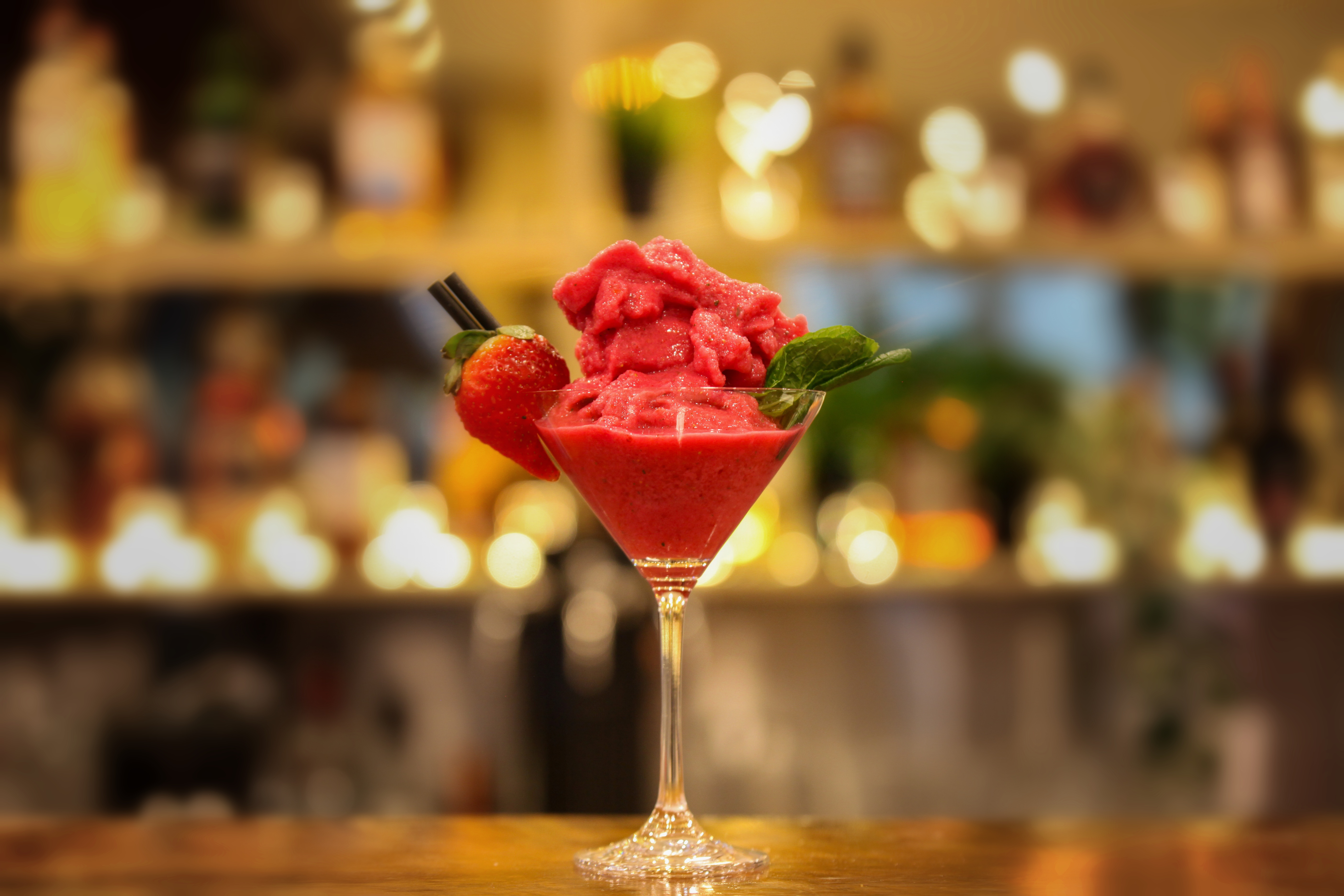 Strawberry daiquiri in a martini glass topped with strawberry sorbet, a fresh strawberry and mint, glowing bar shelves behind