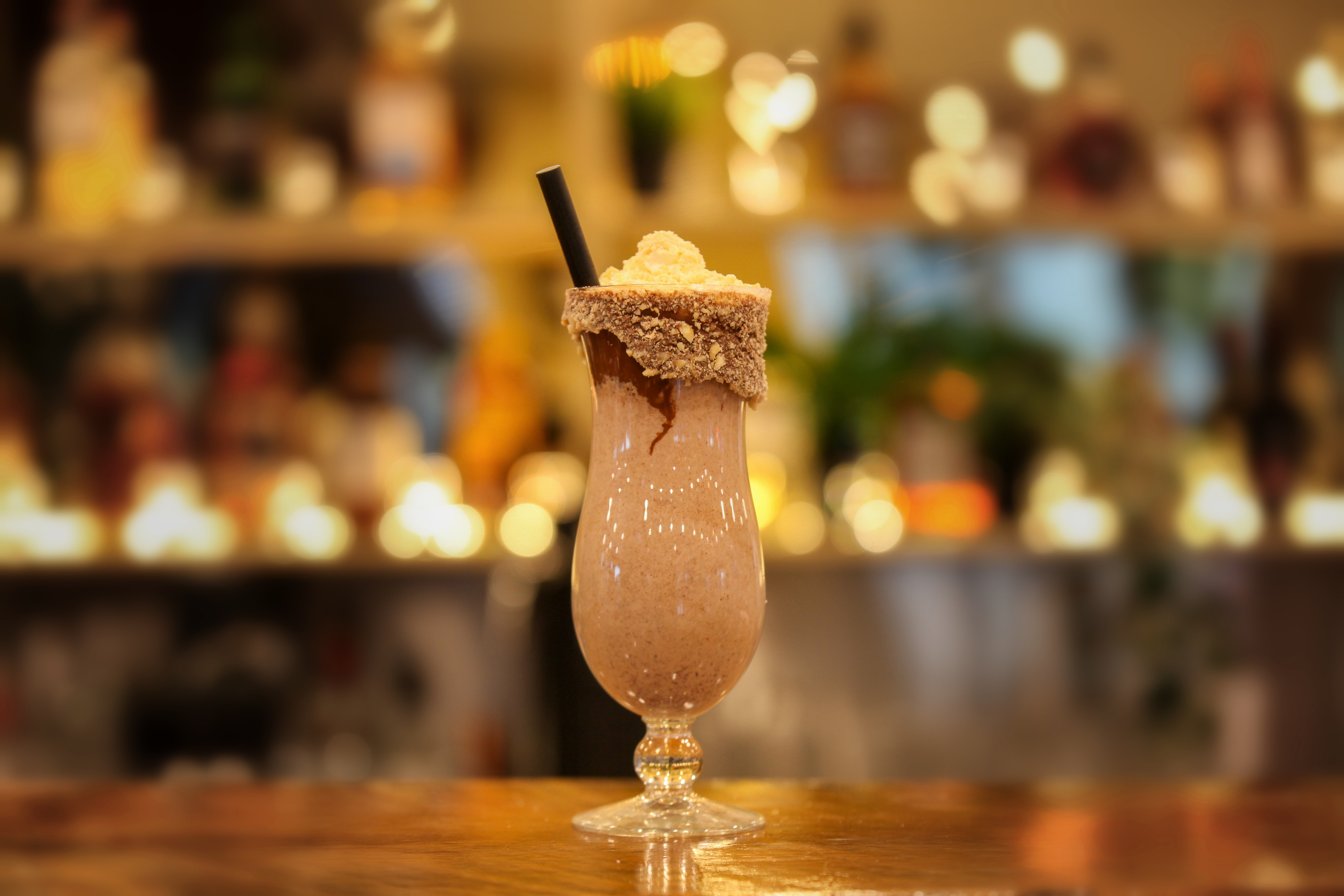 Nutella and hazelnut shake in a hurricane glass with a crushed hazelnut rim, whipped cream and a black straw, glowing bar shelves behind