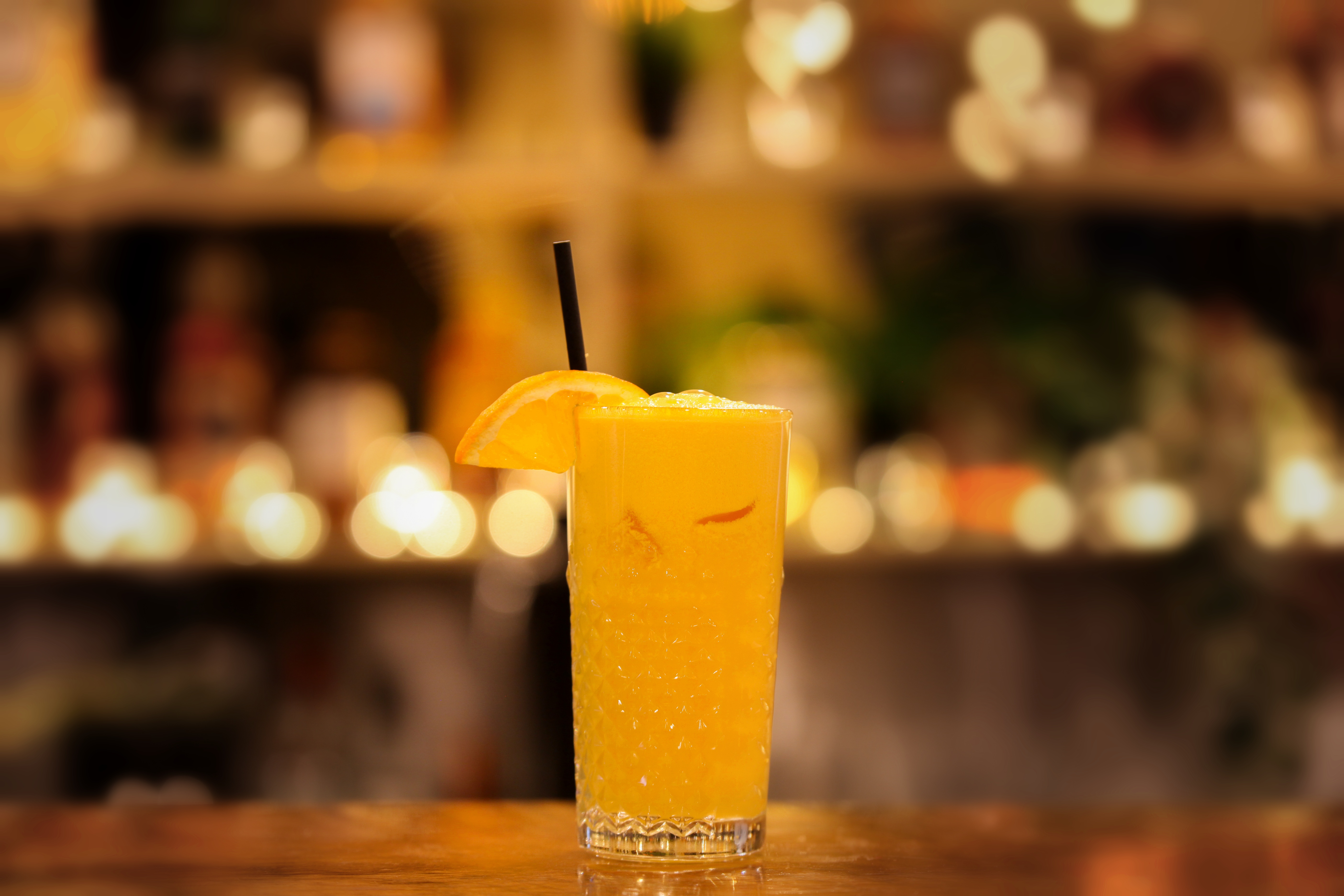 Fresh orange juice in a cut-crystal glass with an orange slice and a black straw, glowing bar shelves behind
