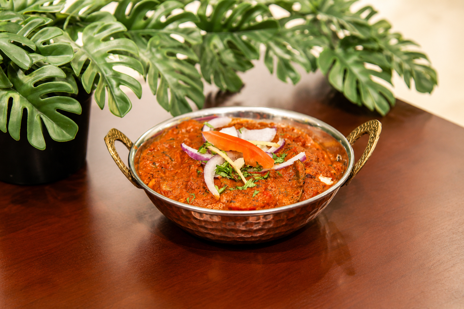 Lamb rogan josh in a hammered copper kadai topped with red onion, tomato, ginger and coriander, monstera plant behind