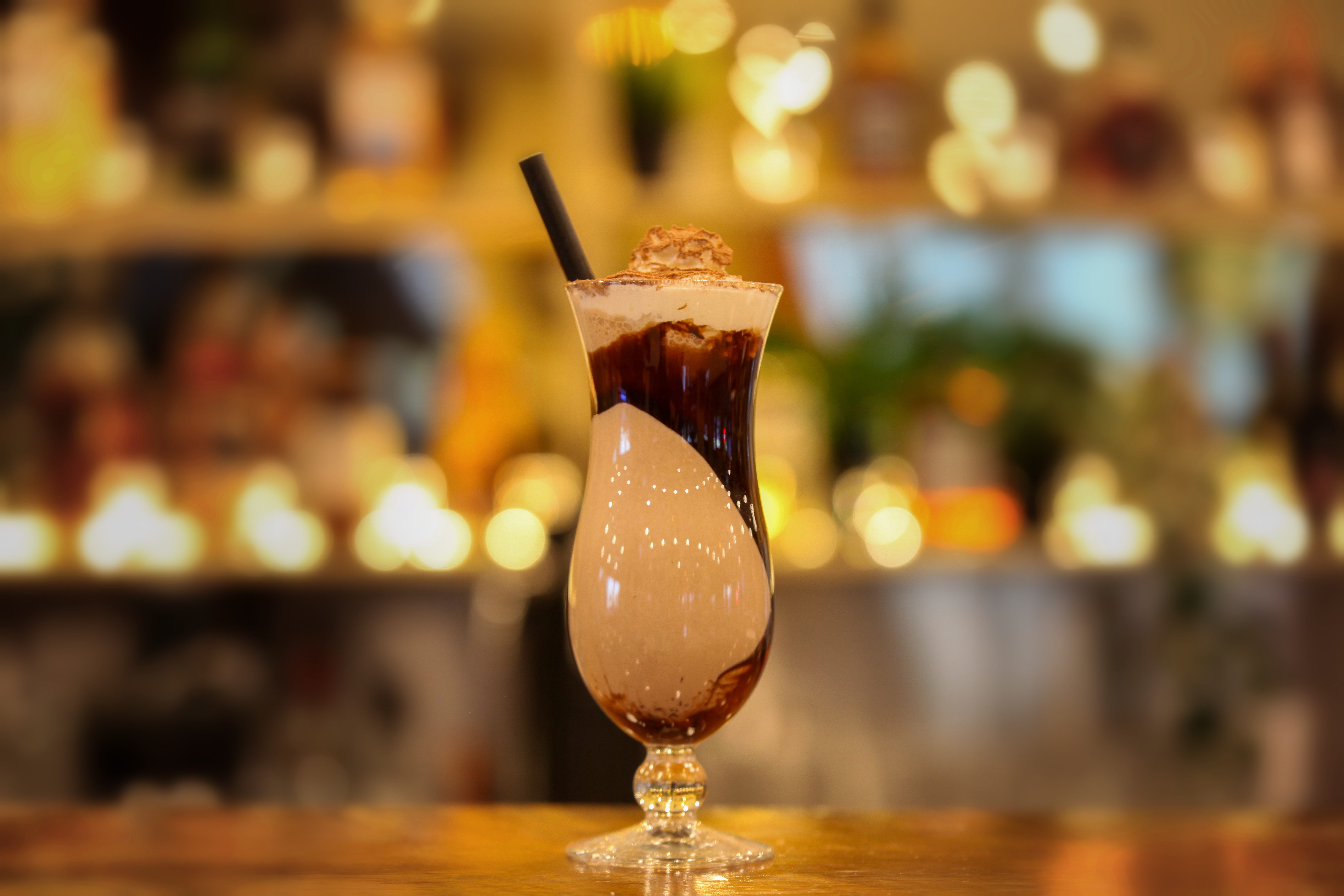 Chocolate shake in a hurricane glass with chocolate sauce swirls, whipped cream and cocoa dusting, glowing bar shelves behind